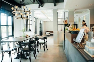 Un interior moderno con comedor que presenta una mesa de madera y sillas negras, una cocina con mostrador que exhibe alimentos, y ventanas amplias que dejan entrar luz natural.