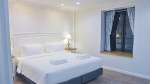 Una cama bien hecha con sábanas blancas, una lámpara de noche y una ventana con cortinas.