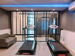 Un gimnasio con dos cintas de correr, un sofá gris y una ventana con persianas.