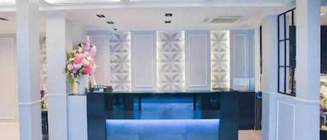Un área de recepción con un mostrador oscuro, paneles decorativos blancos y un jarrón con flores.