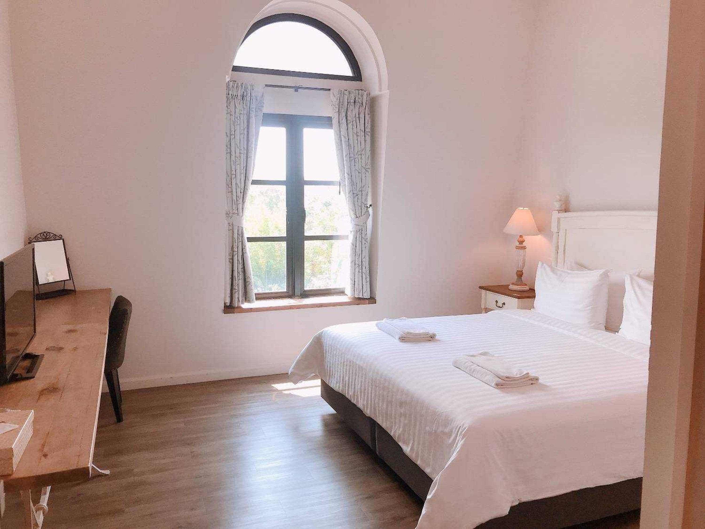 A bedroom with a bed, a desk, a lamp, and a window with curtains.
