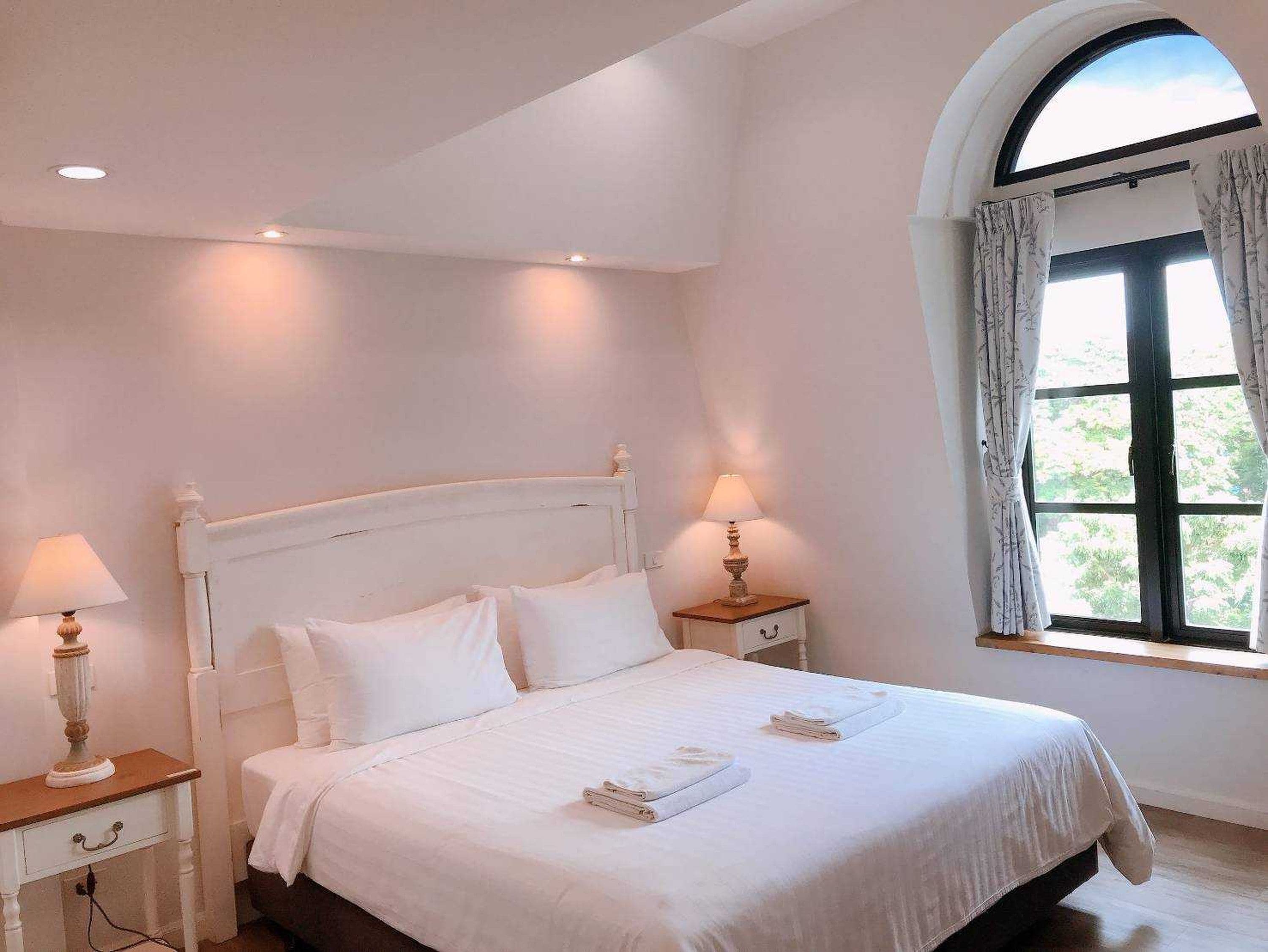 A bedroom with a white bed, two bedside tables with lamps, a window with curtains, and a view of greenery outside.