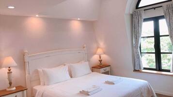 A bedroom with a white bed, two bedside tables with lamps, a window with curtains, and a view of greenery outside.