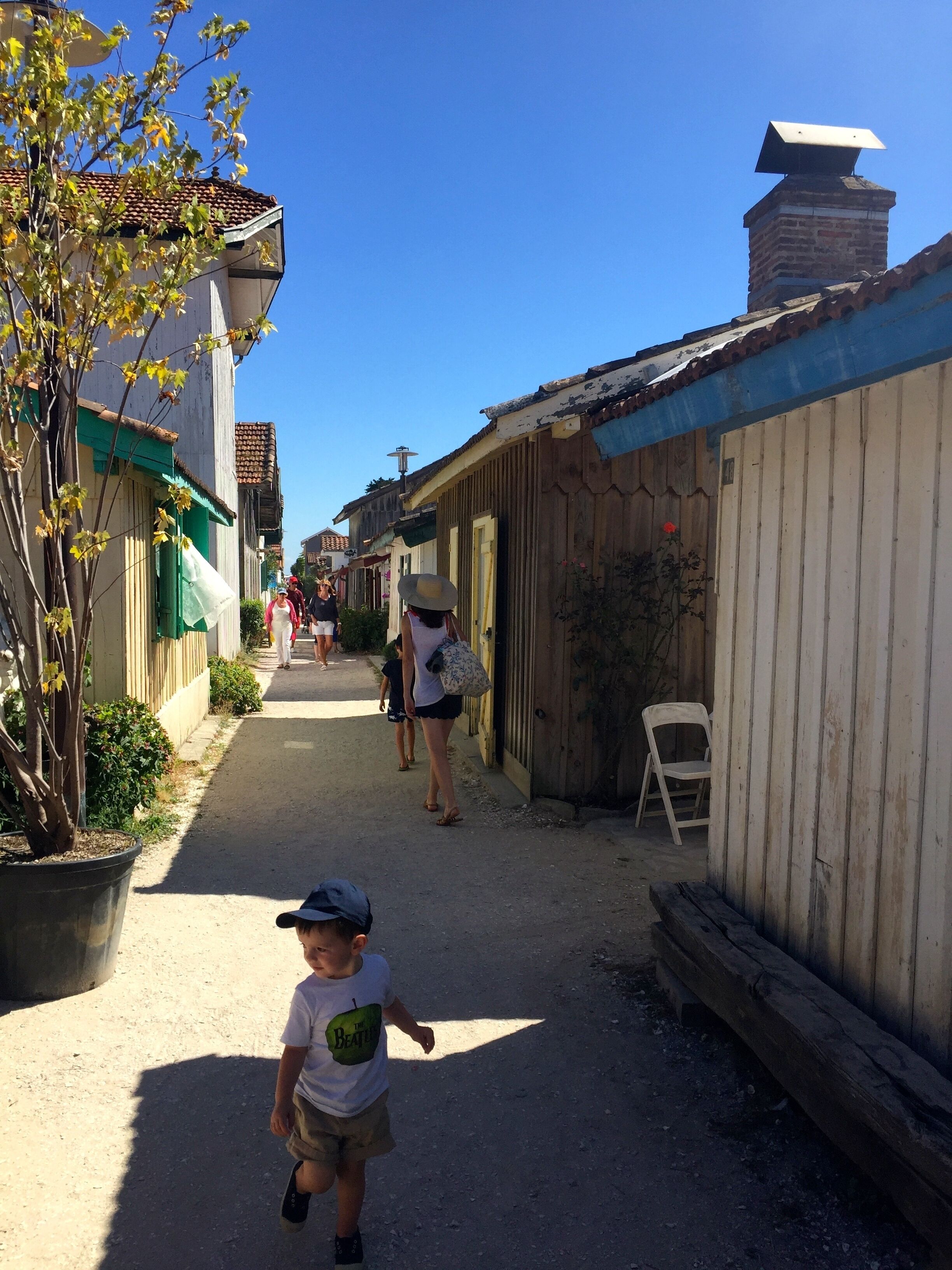 Une rue étroite bordée d’immeubles, un enfant qui marche et une chaise appuyée contre un mur en bois.