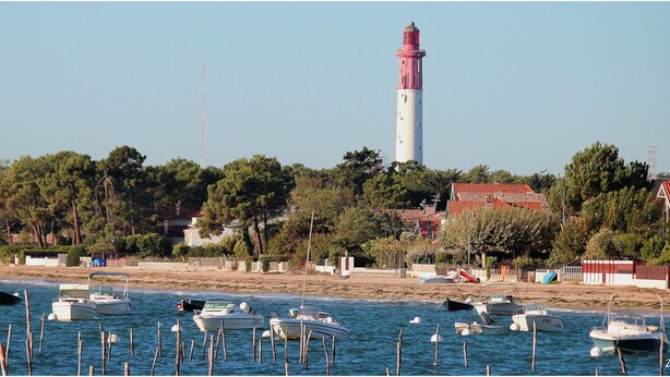 Un paysage côtier avec un phare, des bateaux et une plage bordée de bâtiments.