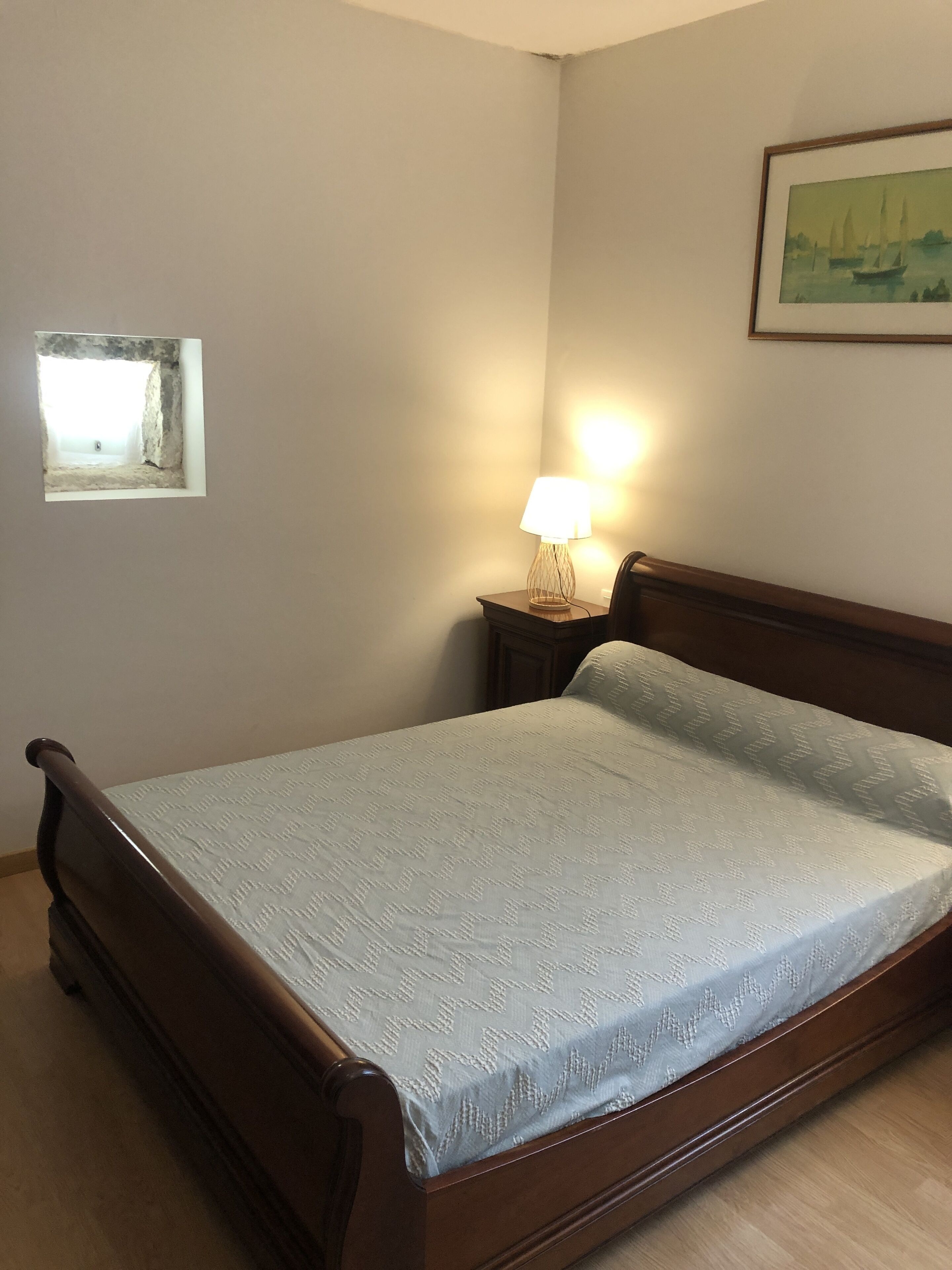 A bedroom with a wooden bed, a nightstand with a lamp, a window, and a painting on the wall.