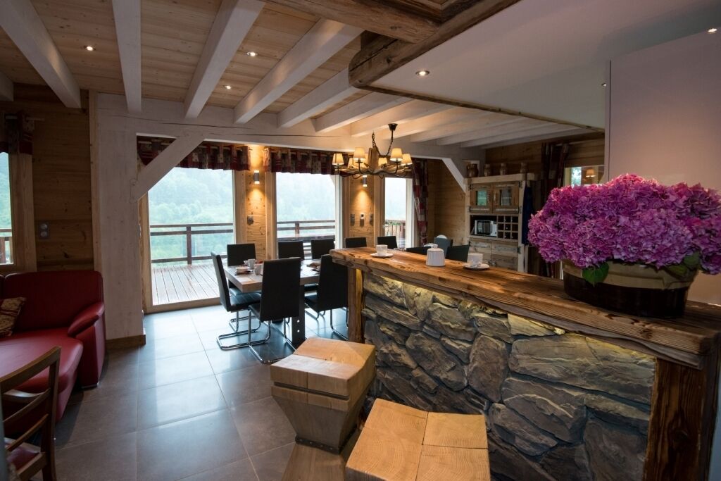 Une pièce spacieuse avec une cheminée en pierre, des poutres apparentes en bois au plafond, un coin repas avec un canapé rouge et une grande fenêtre donnant sur un paysage magnifique.