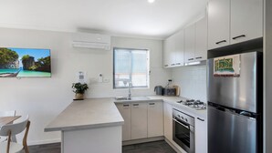 Una cocina moderna con armarios blancos, un refrigerador de acero inoxidable, una estufa a gas y un horno empotrado. Cuenta con una ventana con persianas, un lavamanos y una pequeña isla con una planta.