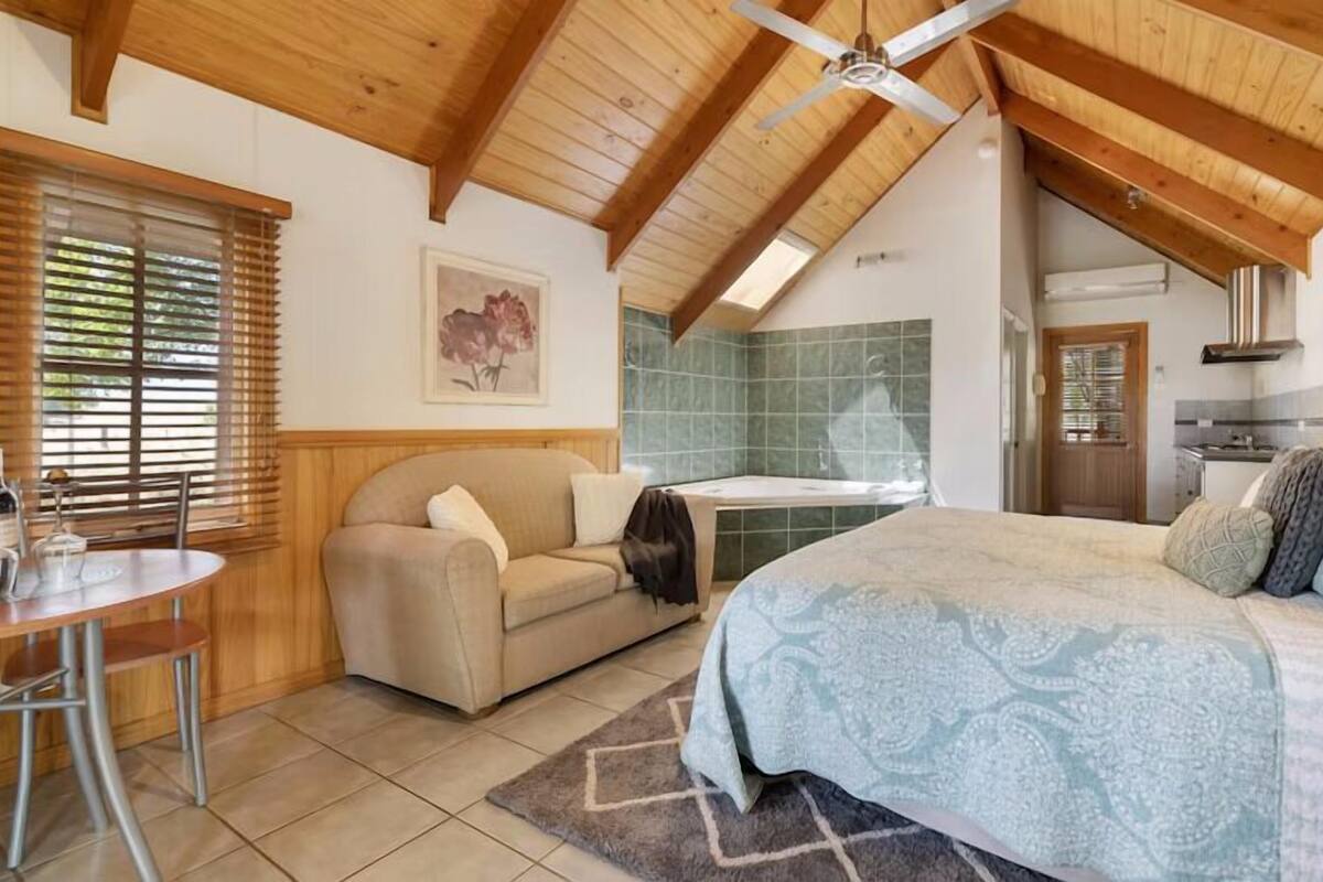A cozy bedroom with a large bed, a small sofa, a dining table, and a kitchen area.