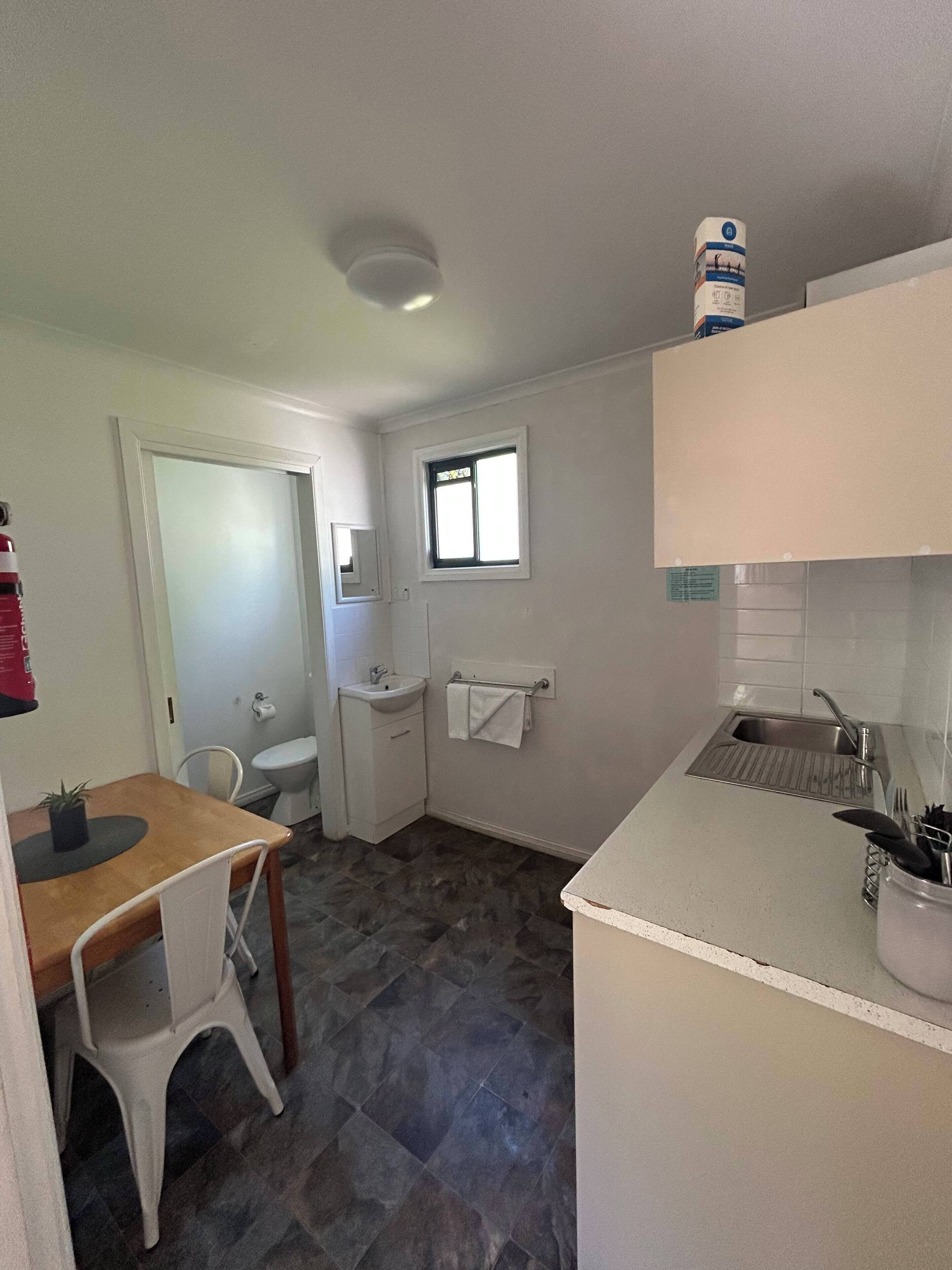 Una cocina compacta con un lavamanos, un ventanal pequeño, una encimera blanca y un comedor con una silla y mesa.