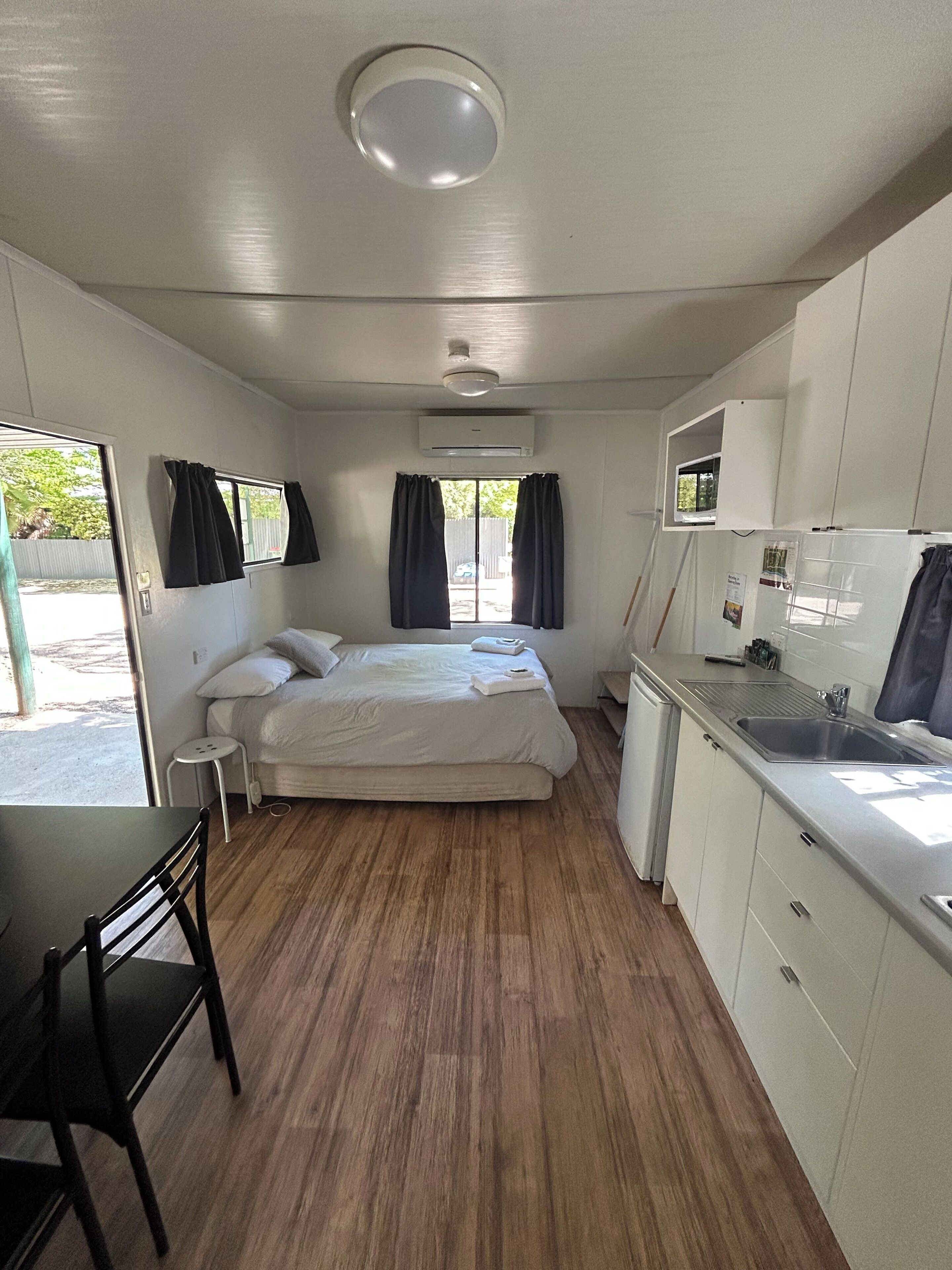 Habitación pequeña con cama, una mesita, una silla, kitchenette y ventana con cortinas.