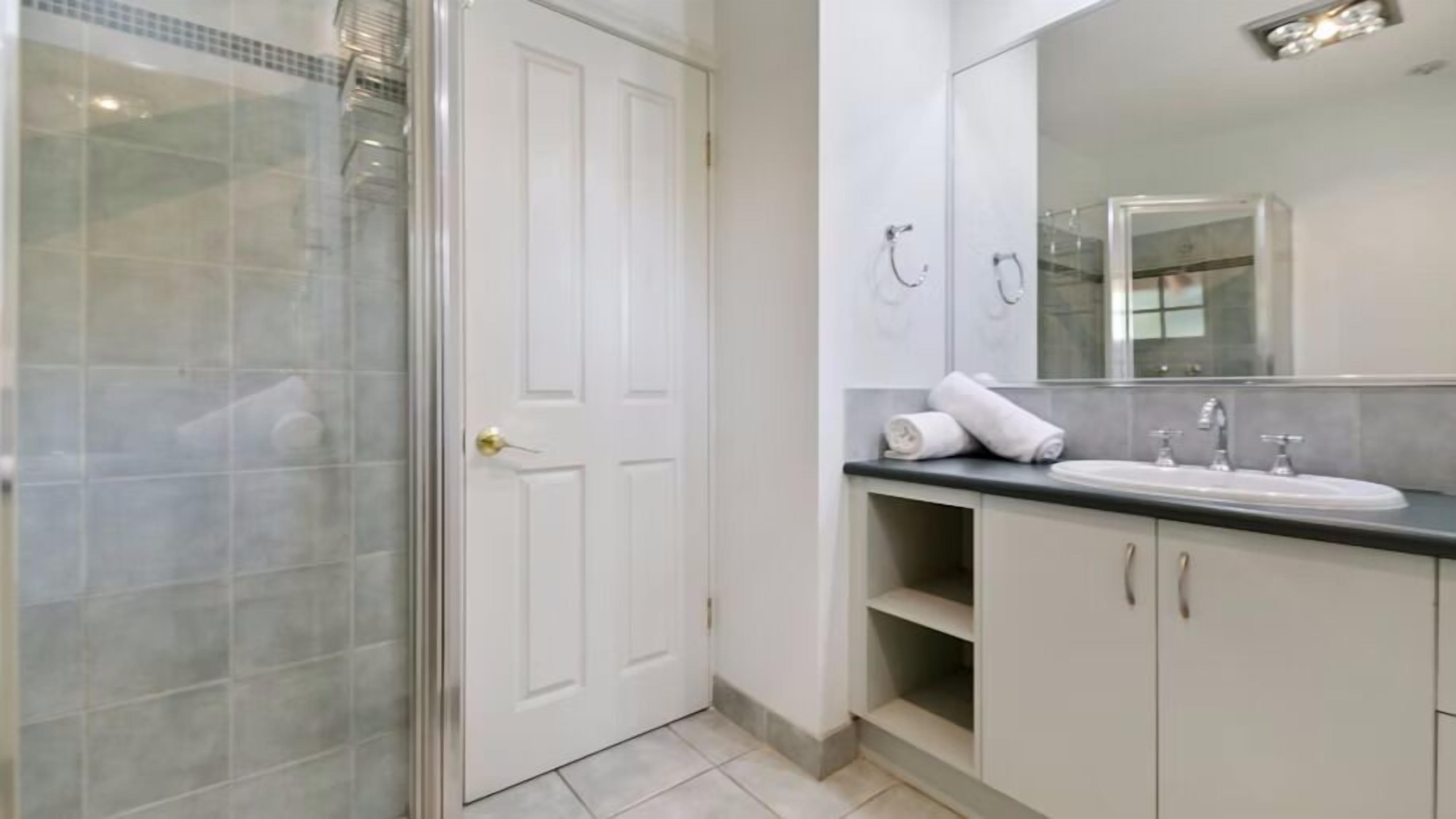 Un baño con cabina de ducha de vidrio, una puerta blanca, un lavabo con dos grifos y un espejo sobre el lavabo.