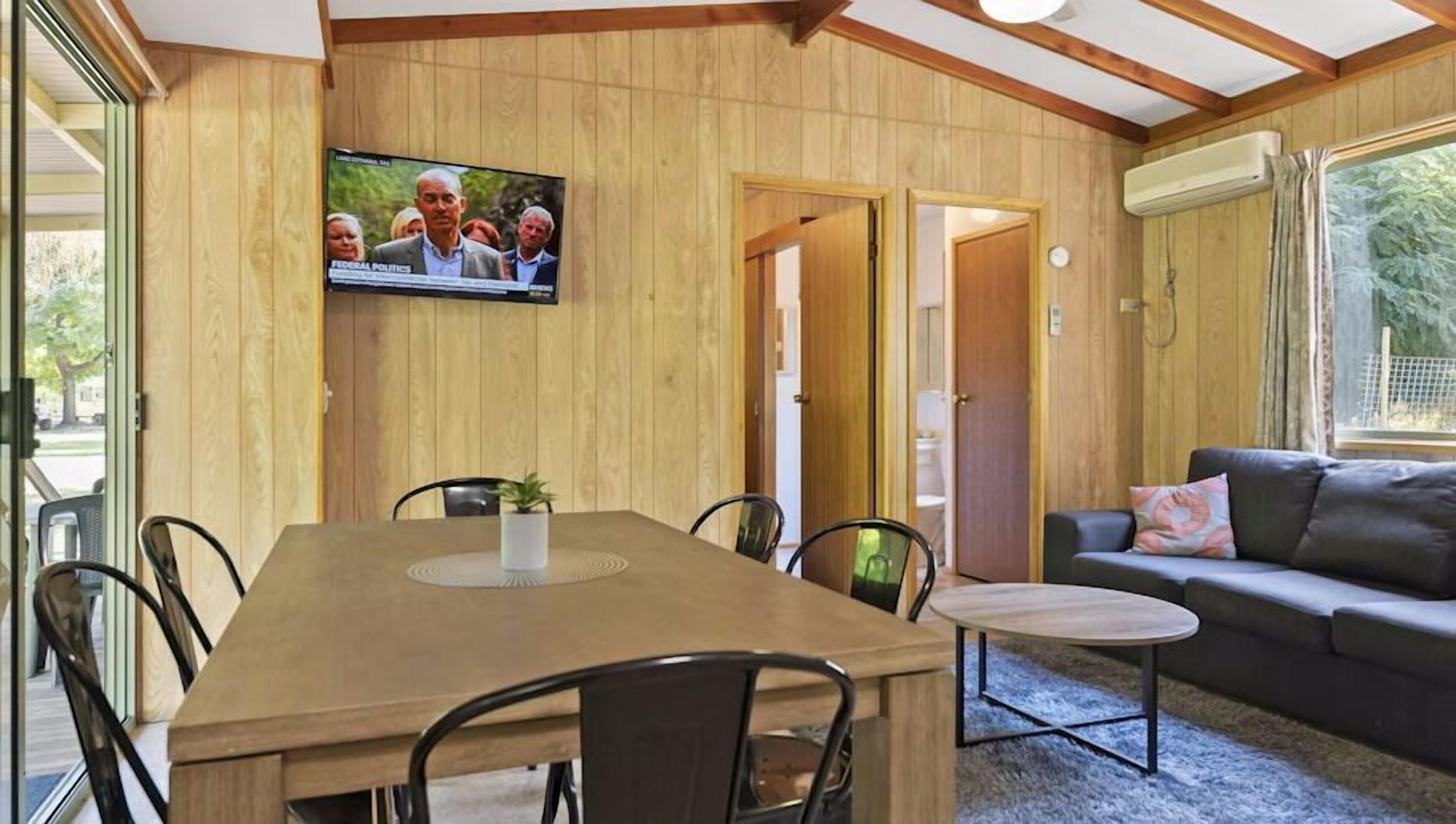 Ein Zimmer mit einer holzverkleideten Wand, einem Fernseher über einem Esstisch mit Stühlen, einer Couch und einem kleinen runden Tisch.