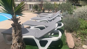 Un espace aménagé au bord de la piscine, avec des chaises longues et un parasol, entouré de verdure et bordé d’un bâtiment dont le toit est recouvert de tuiles.
