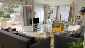 Un salon moderne avec des canapés gris, un fauteuil jaune et une table basse en verre. On y trouve un téléviseur posé sur un meuble, un coin repas avec des chaises blanches et un balcon agrémenté de plantes.