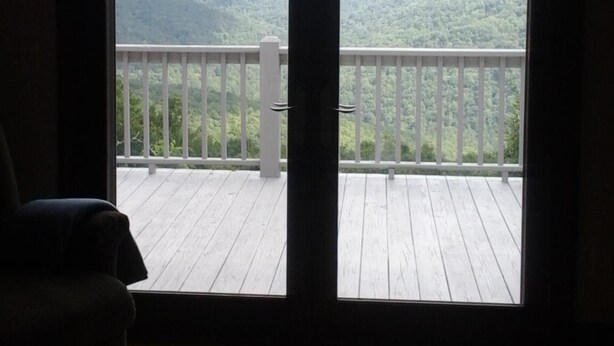 Ein Zimmer mit einer großen Schiebeglastür, die auf einen Balkon mit Holzdeck und Blick auf eine bewaldete Landschaft führt.