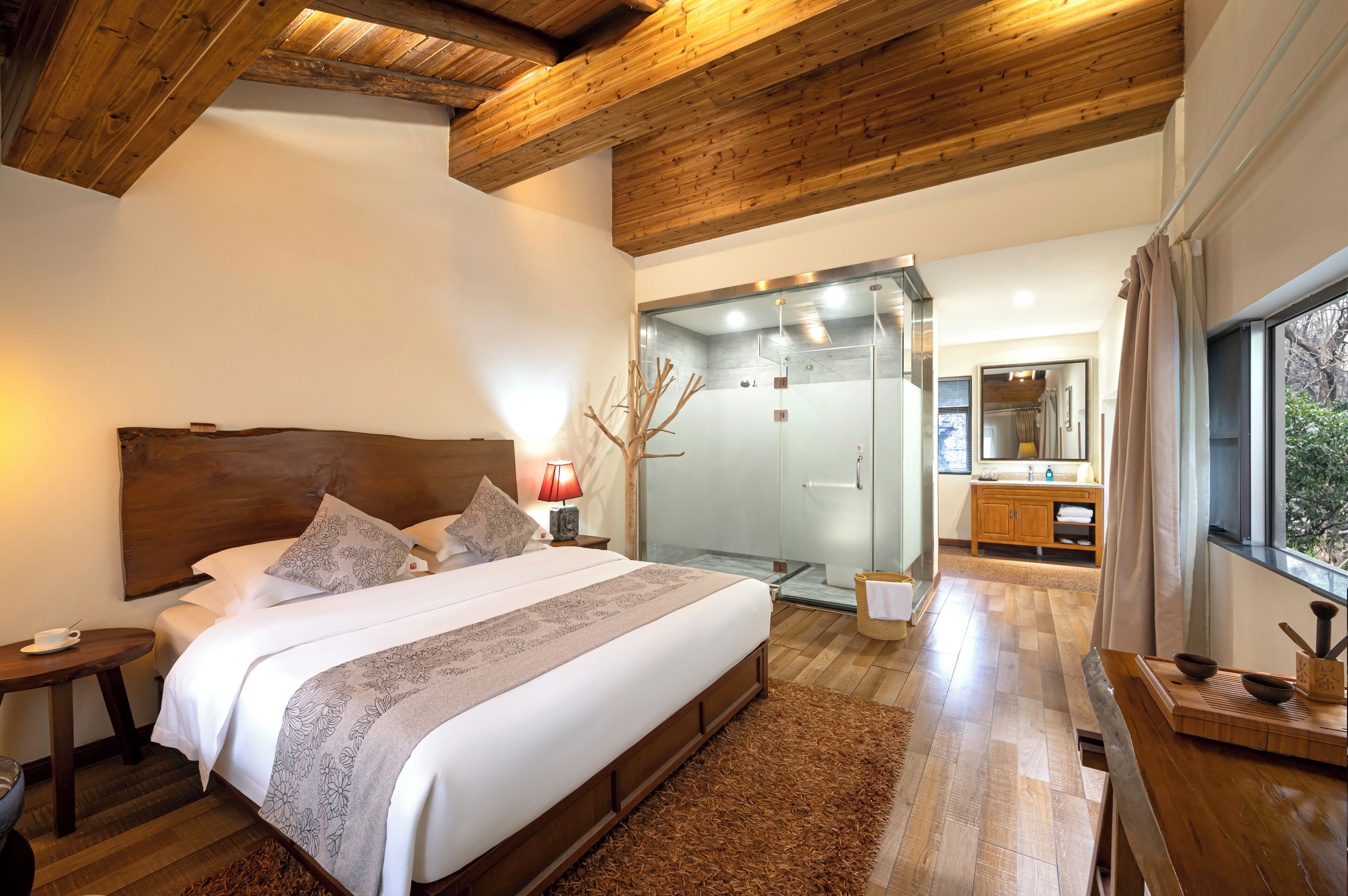 A modern bedroom with a large bed, wooden ceiling beams, a glass shower, and a view of the outdoors.