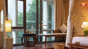 A room with a wooden bench, a bedside table with a lamp, a wicker basket, and a view of greenery through large windows. - Guilin Crystal Crescent Moon Hotel (Guilin)