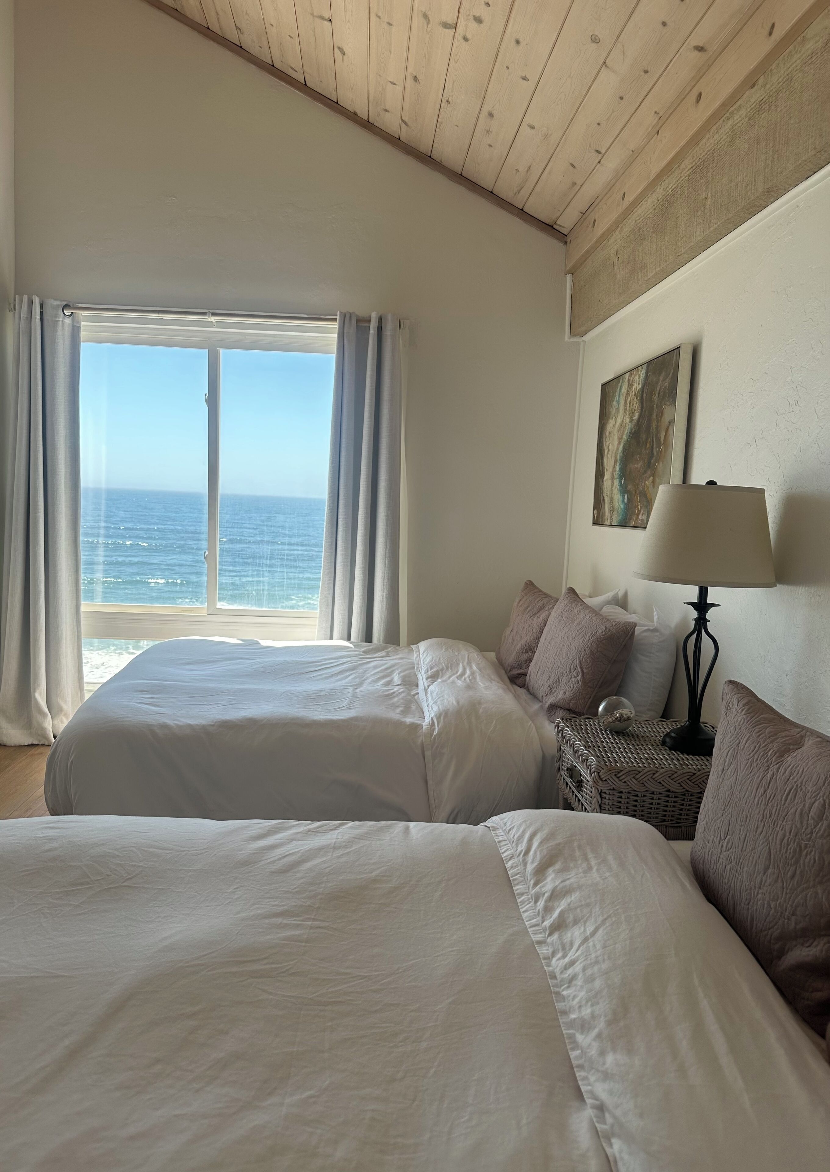 A bedroom with two beds, a view of the sea, a painting on the wall, and a lamp on a side table.