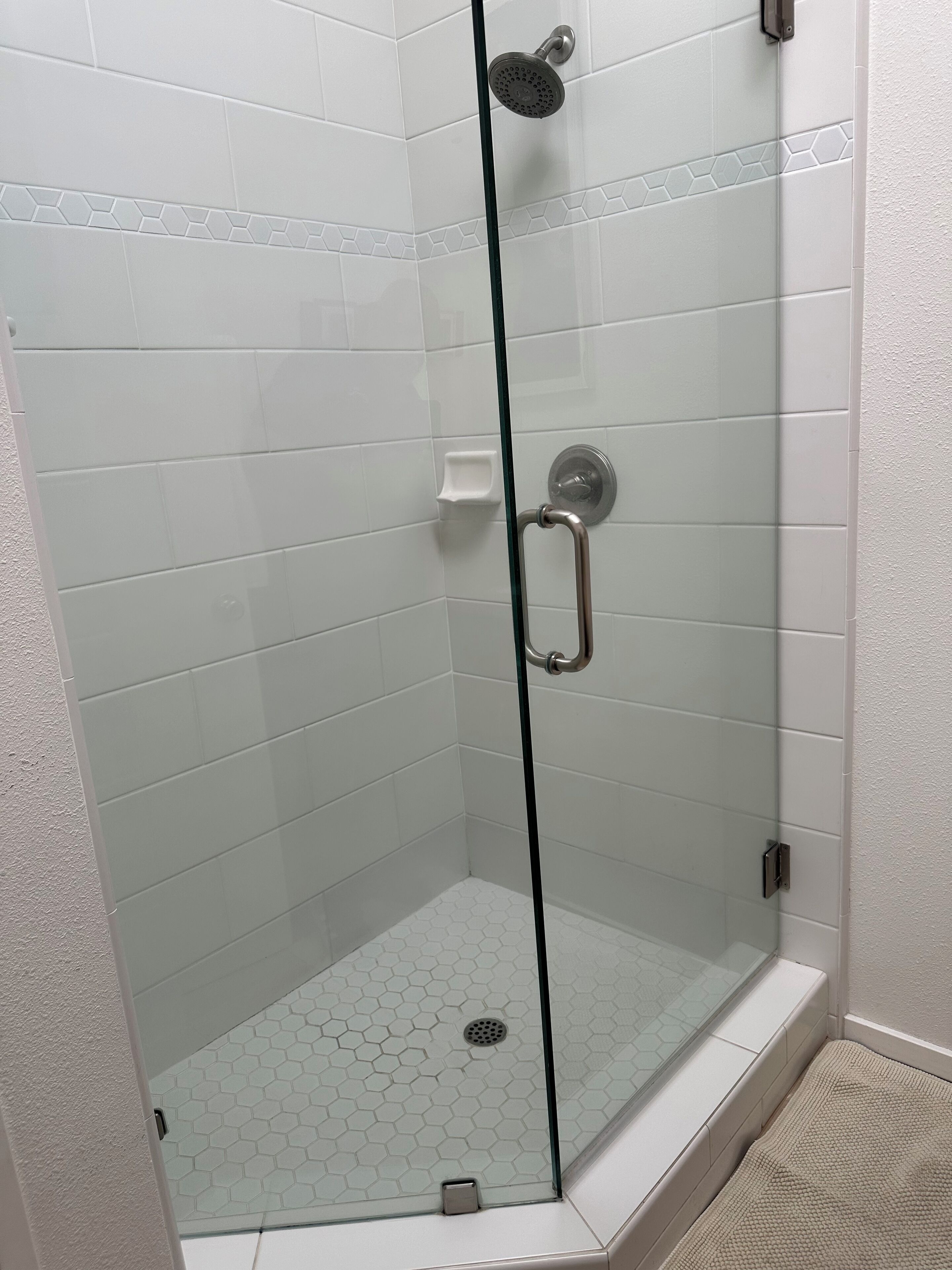 Eine begehbare Dusche mit Glastrennwand, weißen sechseckigen Fliesen, einer Handbrause und einer Glastür mit Griff.
