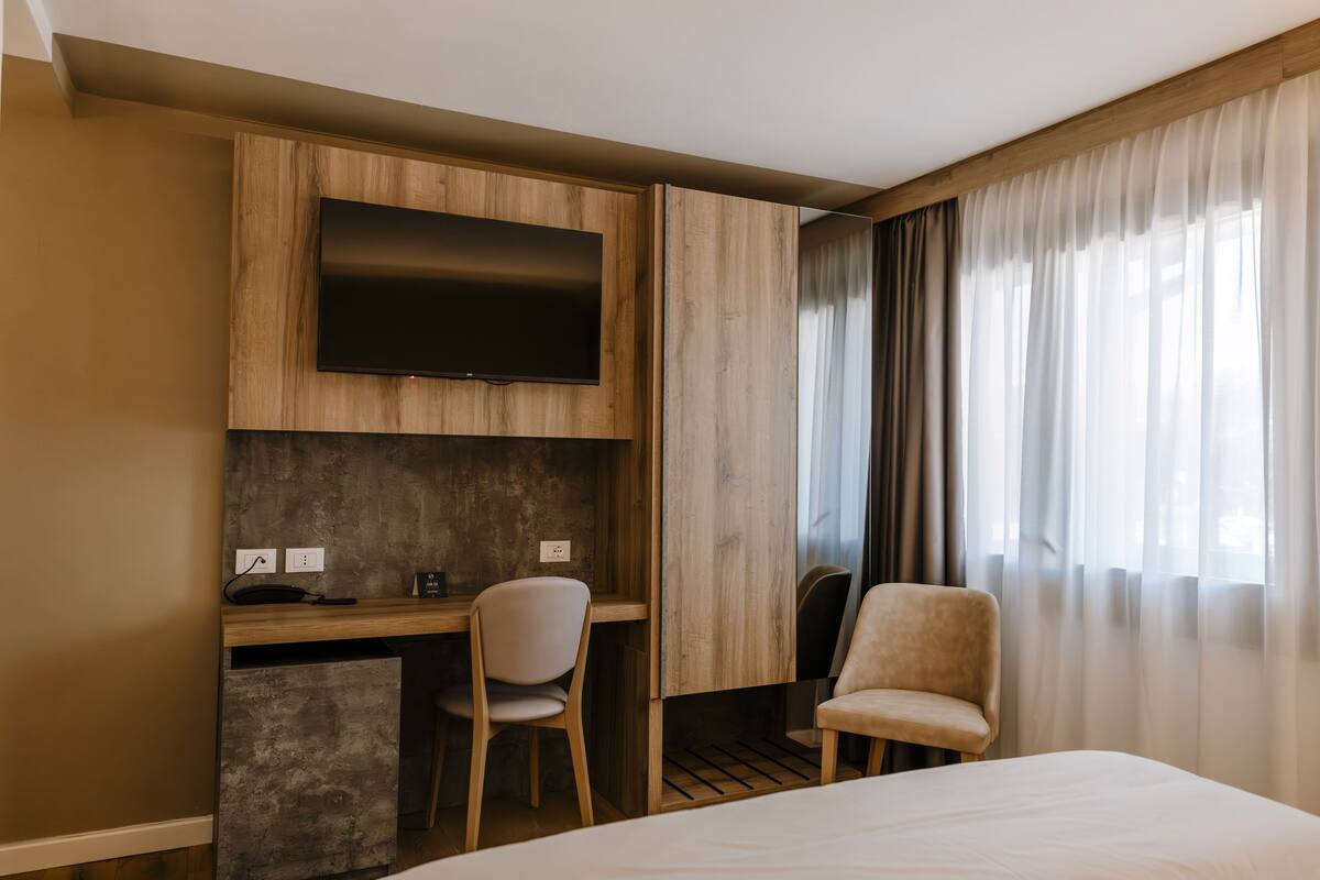 A hotel room with a bed, a desk with a chair, a television mounted on the wall, and a window with curtains.