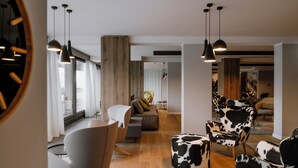 A modern living room with a sofa, chairs, and a round ottoman. There are pendant lights and a large window with curtains. - Armon Greenblu Hotel & Wellness (Dimaro Folgarida)
