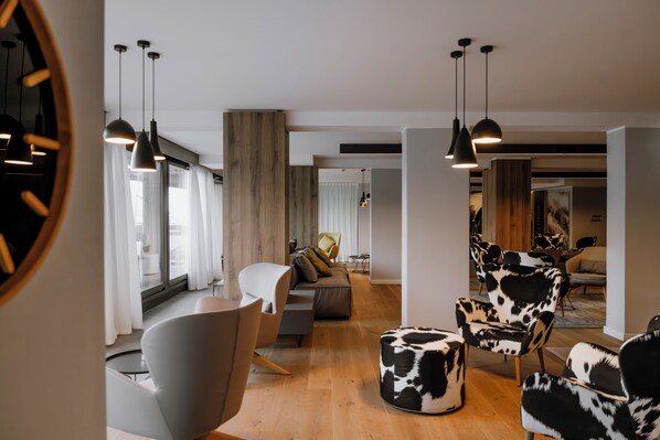 A modern living room with a sofa, chairs, and a round ottoman. There are pendant lights and a large window with curtains. - Armon Greenblu Hotel & Wellness (Dimaro Folgarida)