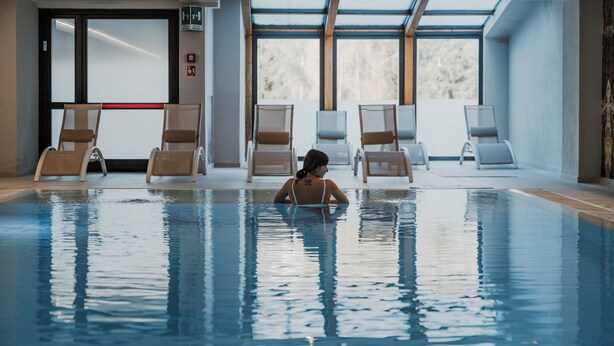 An indoor swimming pool with clear glass walls, multiple lounge chairs, and a visible exit sign. - Armon Greenblu Hotel & Wellness (Dimaro Folgarida)