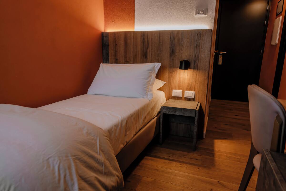 A hotel room with a wooden headboard, a single bed with white bedding, a nightstand with a lamp, and a wooden floor.