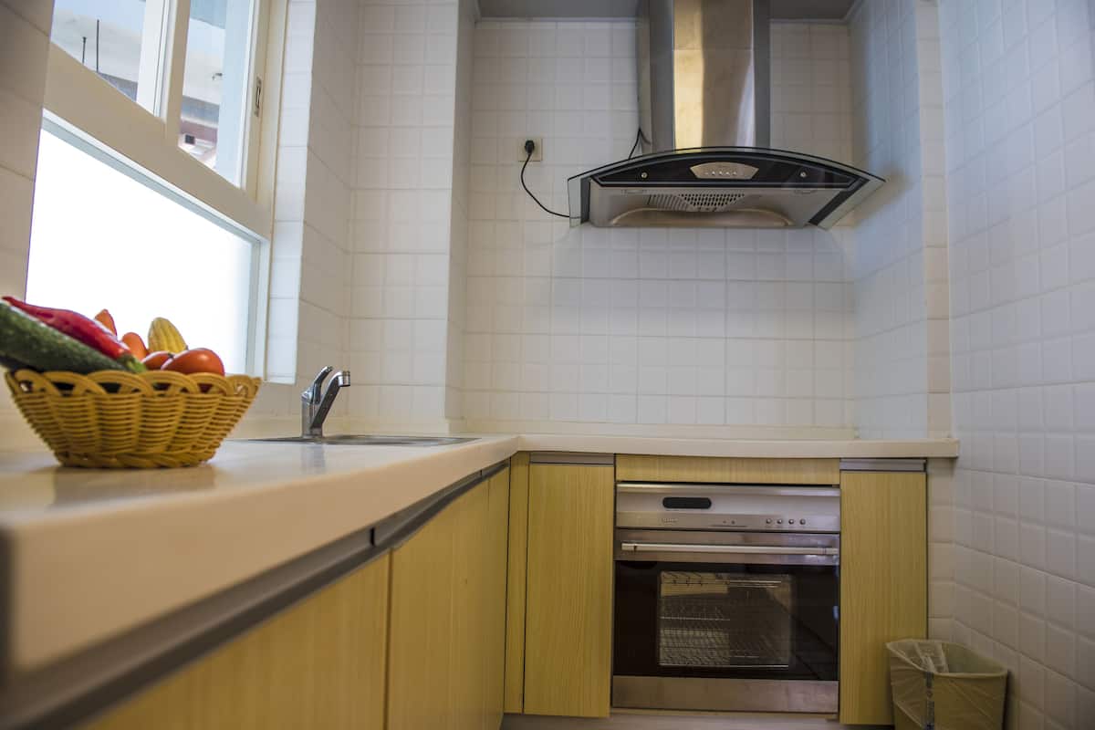 A kitchen with white tiled walls, a stainless steel range hood, a built-in oven, and a basket of fruits on the counter.