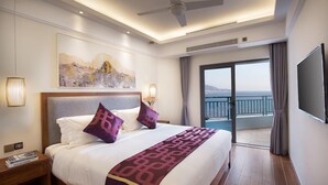 Habitación de hotel con cama, un cuadro en la pared, ventilador de techo y vista al mar desde el balcón.