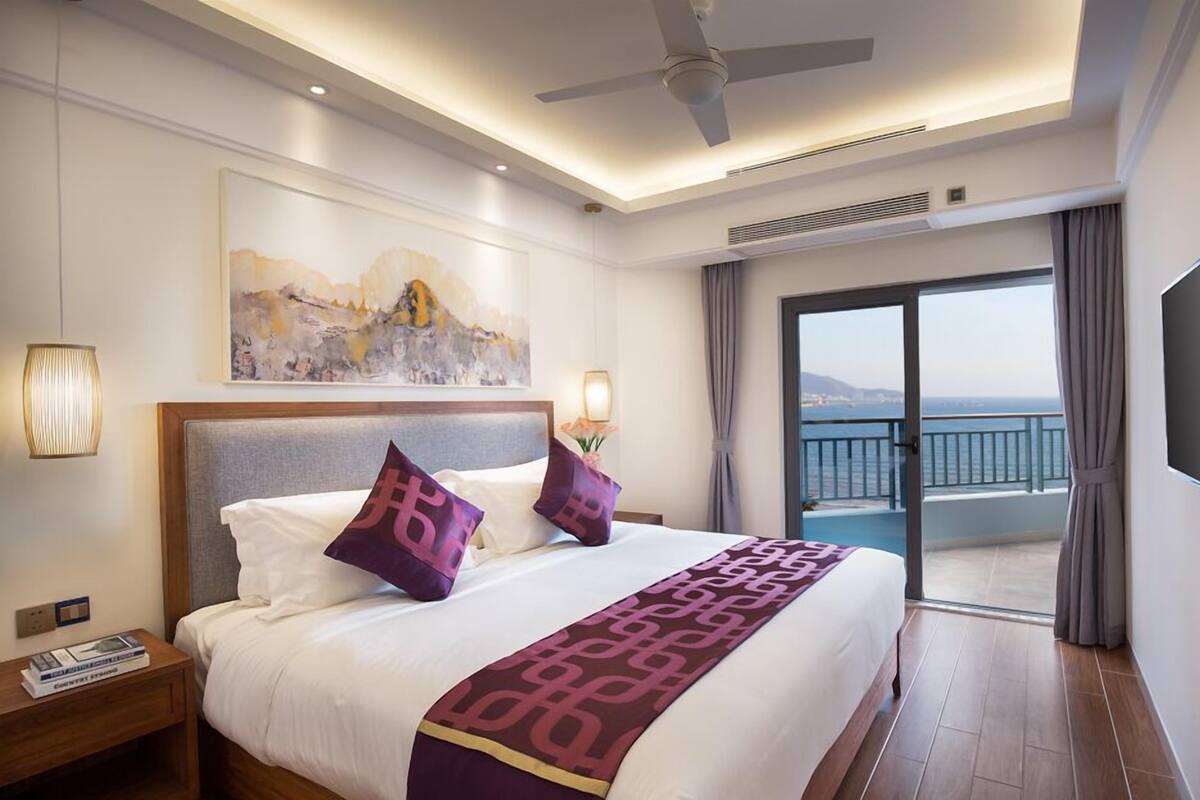 A hotel room with a bed, a painting on the wall, a ceiling fan, and a view of the sea through a balcony.