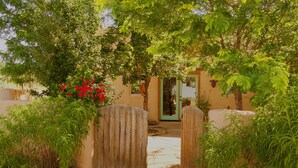 A wooden gate leading to a house with a green door, surrounded by lush greenery and a gravel path.