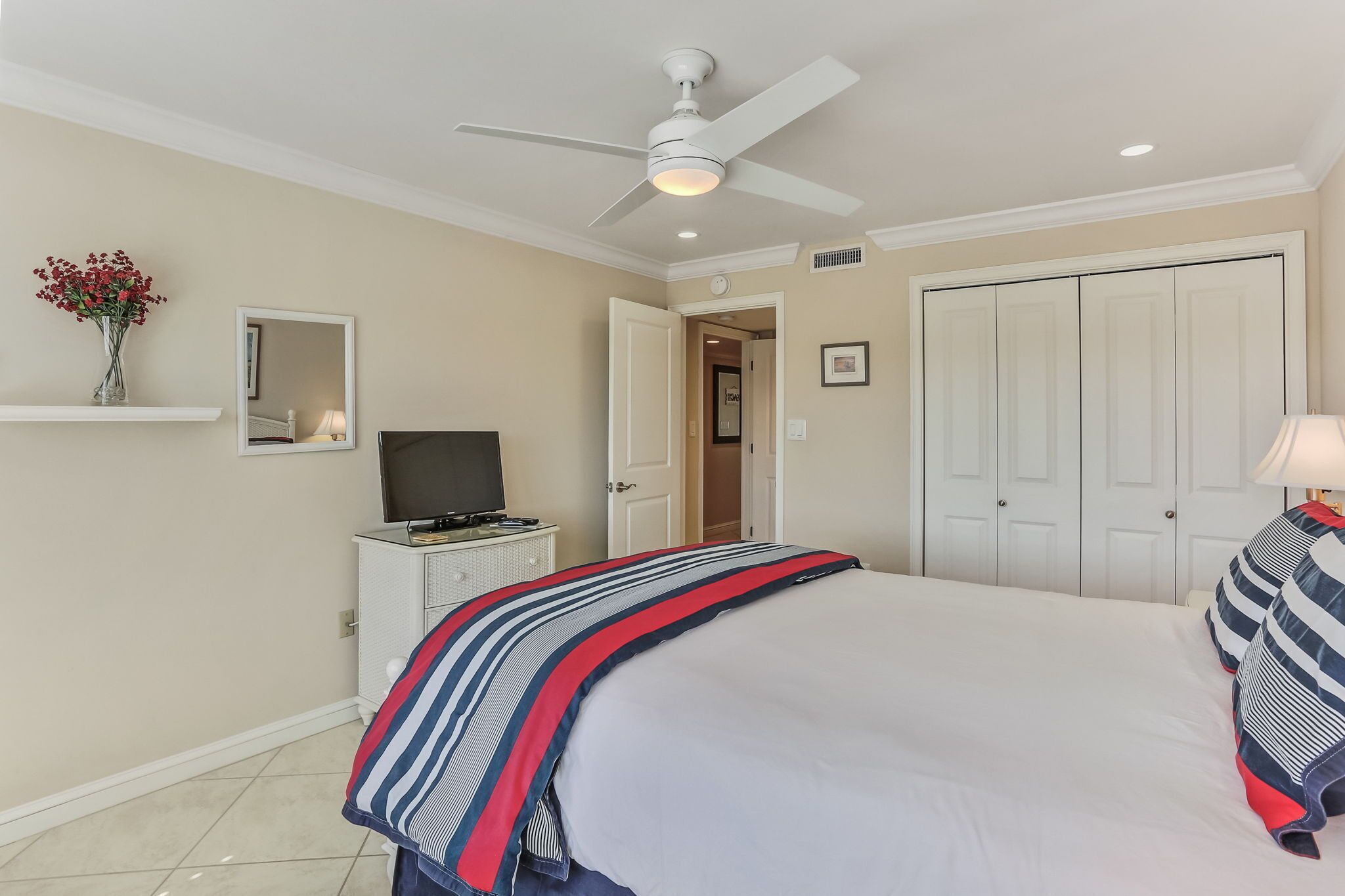 A bedroom with a bed, a ceiling fan, a TV, a dresser, a mirror, and a door leading to another room.