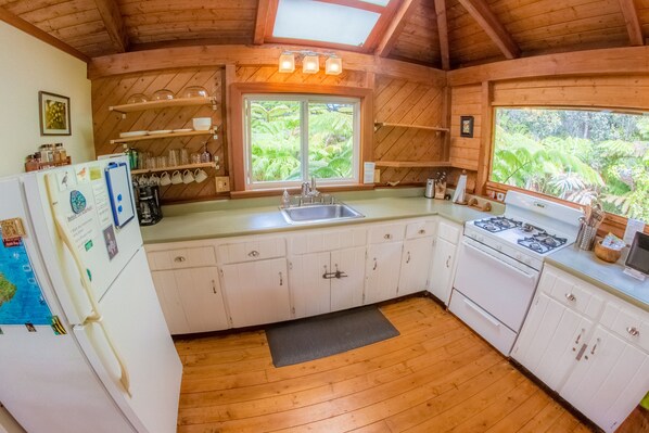 A kitchen with wooden cabinets, a sink, and a stove. There are shelves above the sink and a window with a view of greenery. - 8 minutes to Nat'l Park! Enjoy the sound of rain & birds in a peaceful retreat. (Volcano)