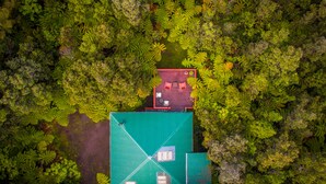 An aerial view of a house surrounded by dense forest. The house has a green roof and a red deck area with two chairs and a table. - 8 minutes to Nat'l Park! Enjoy the sound of rain & birds in a peaceful retreat. (Volcano)