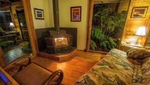 A cozy room with a fireplace, a comfortable chair, a sofa with a floral pattern, and a view of tropical plants through a glass door. - 8 minutes to Nat'l Park! Enjoy the sound of rain & birds in a peaceful retreat. (Volcano)
