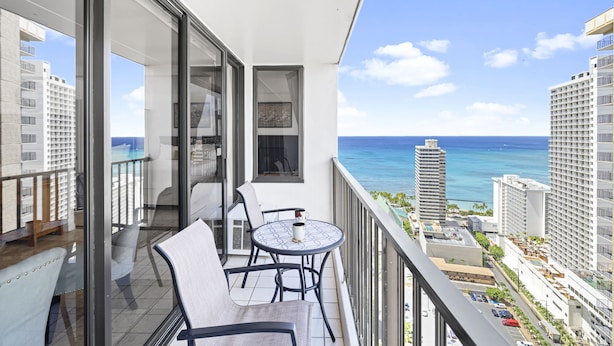 A balcony with a table and chairs, overlooking a cityscape and the ocean. - High Rise Deluxe Ocean View Free Parking/Wifi in Waikiki 3204 (Honolulu)