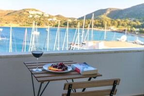 A wooden table with a glass of red wine, a plate of fruit, and a book. The table is on a balcony with a view of a marina and hills in the background. - Bora Hotel (Datça)