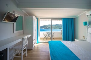 Habitación de hotel con cama, escritorio con silla, televisión y vista al mar a través de una puerta corrediza de vidrio.