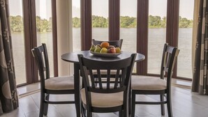 Una mesa de comedor con sillas y un frutero, frente a una vista de agua y vegetación.