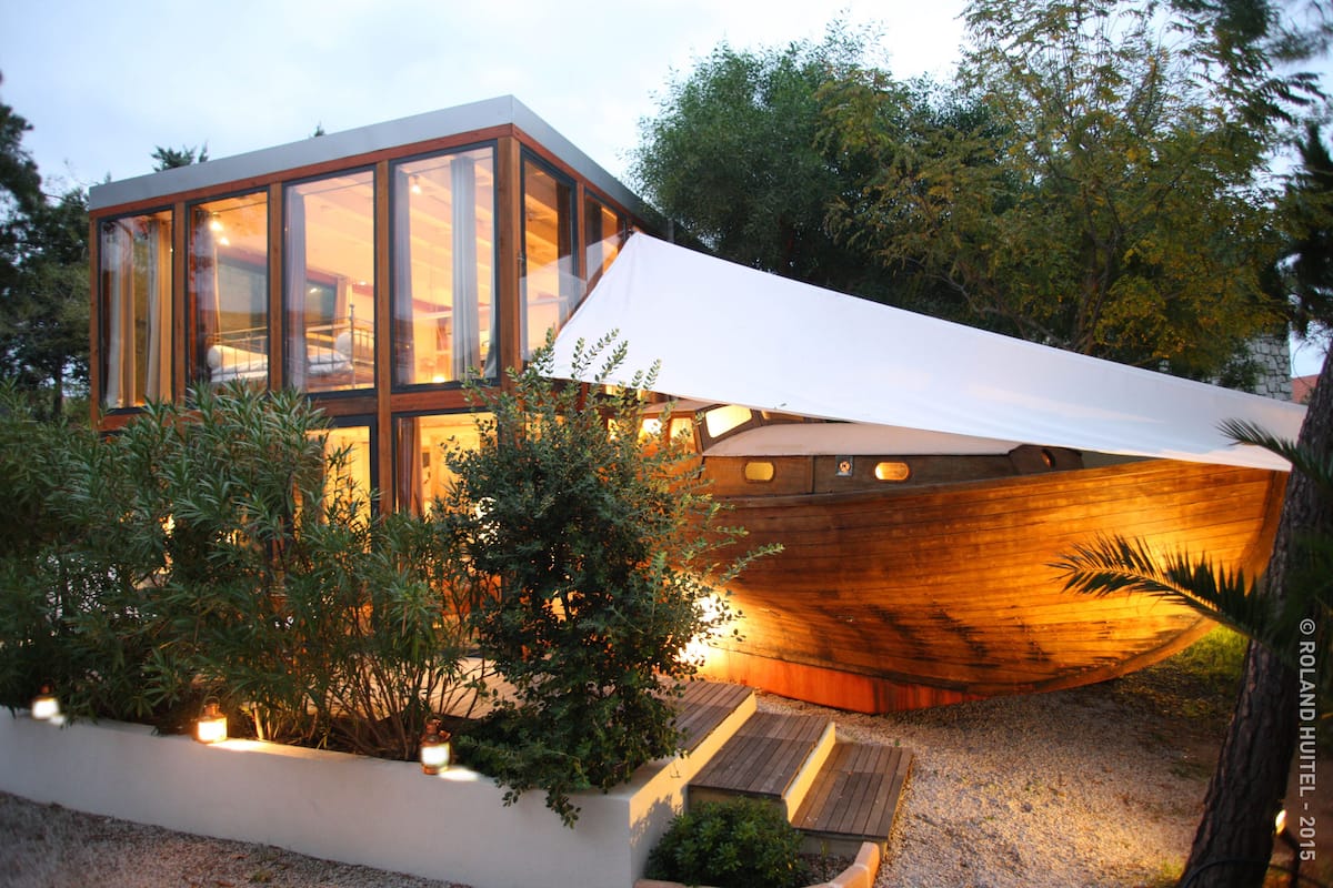 A modern house with large glass windows, a wooden boat-shaped extension, and a garden with plants and a pathway.