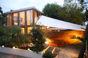 Ein modernes Haus mit großen Glasfenstern, einer bootförmigen Holzkonstruktion und einem Garten mit Pflanzen und einem Weg.