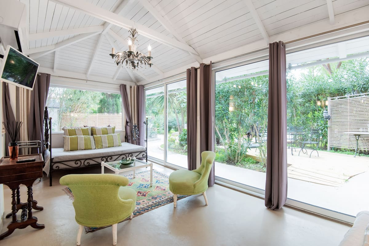 A spacious living room with a large window, a sofa, two green armchairs, and a chandelier.