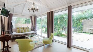 A spacious living room with a large window, a sofa, two green armchairs, and a chandelier.