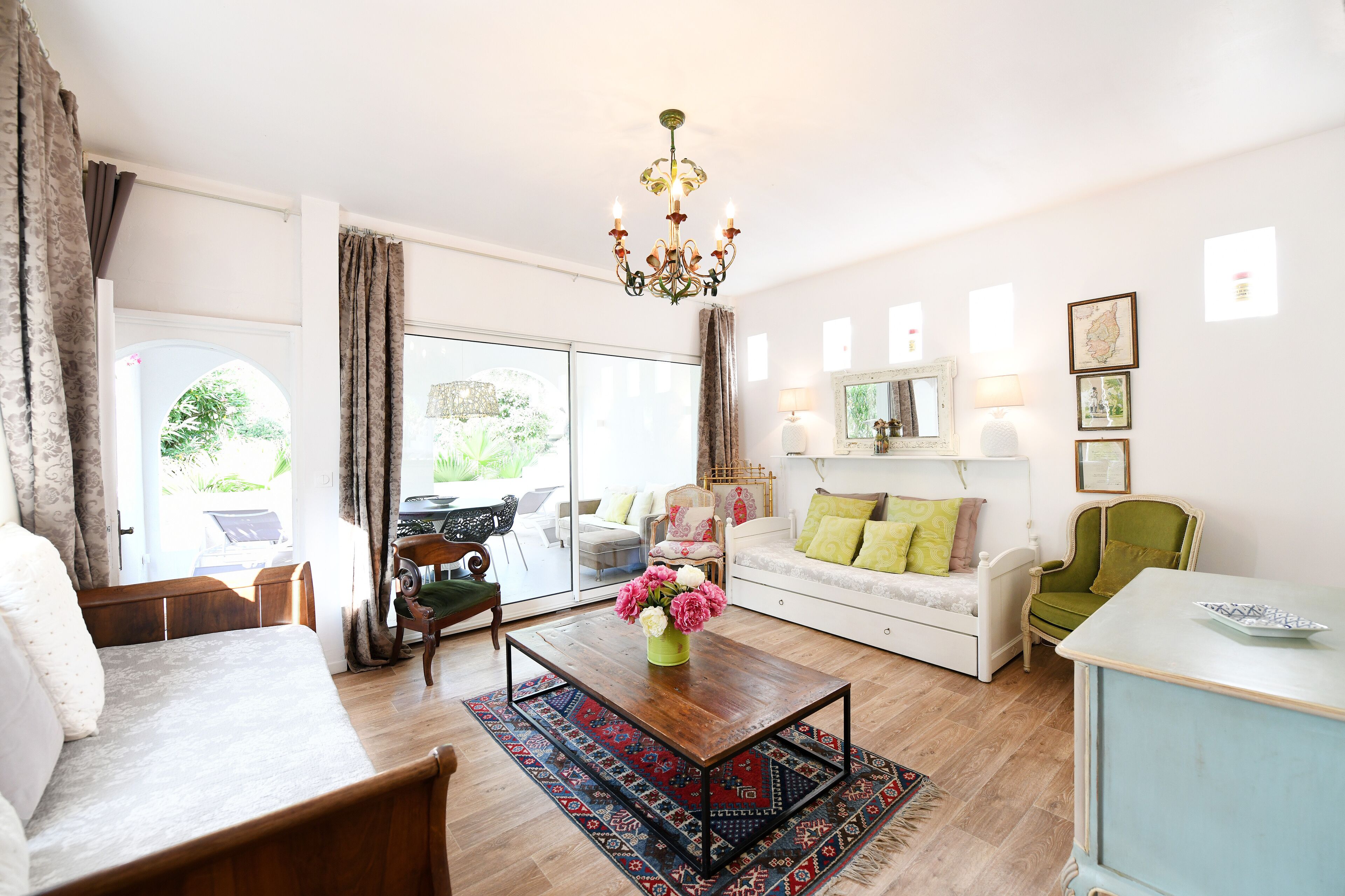 Un salon spacieux avec une table basse en bois, un canapé, un fauteuil vert et un fauteuil blanc. Un lustre est suspendu au plafond, et une grande fenêtre est agrémentée de rideaux.
