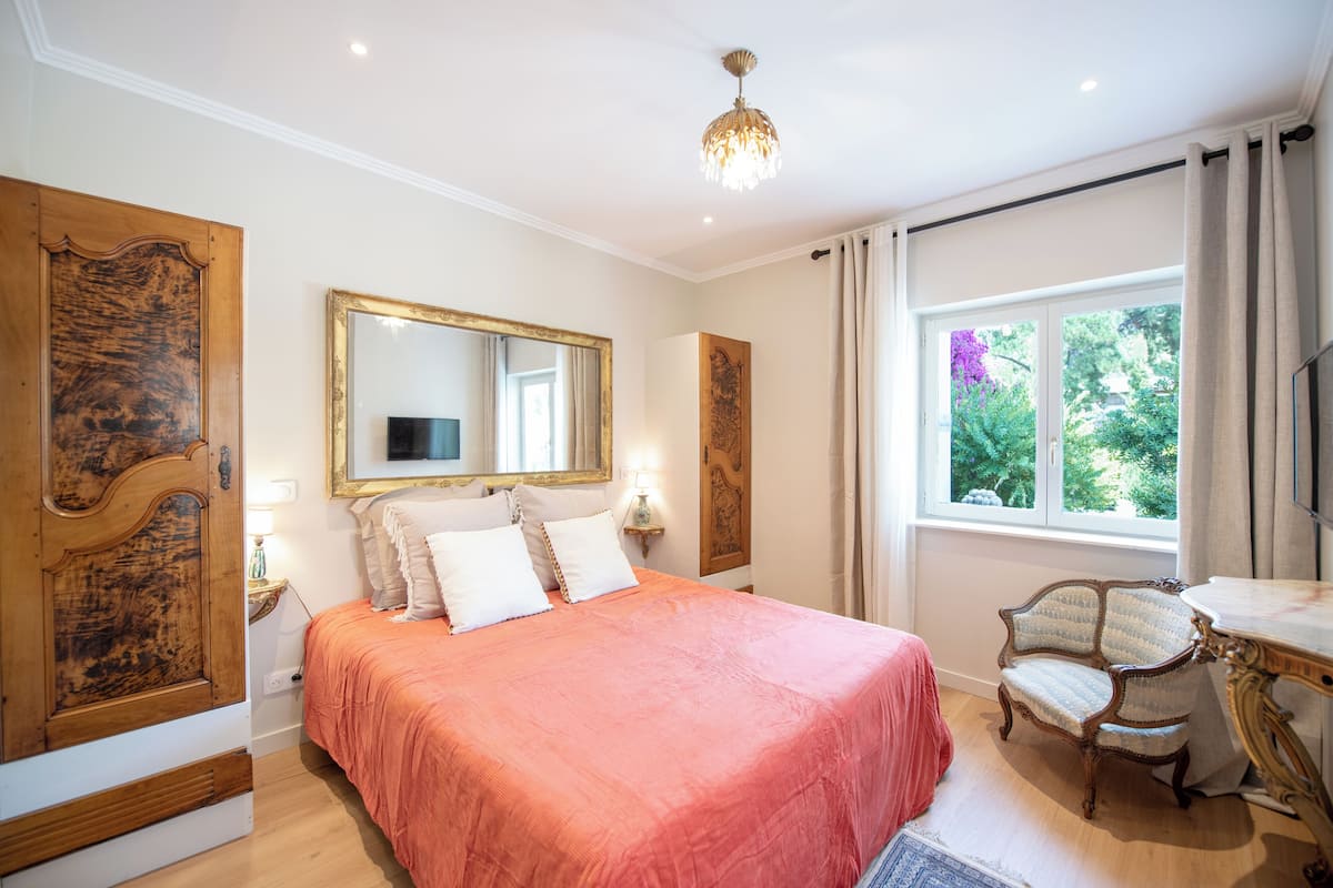 A bedroom with a large bed, a wooden chair, a mirror, and a window with curtains.