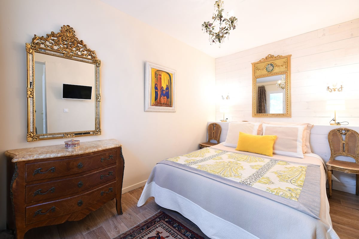A bedroom with a bed, dresser, two mirrors, a painting, and a chandelier.