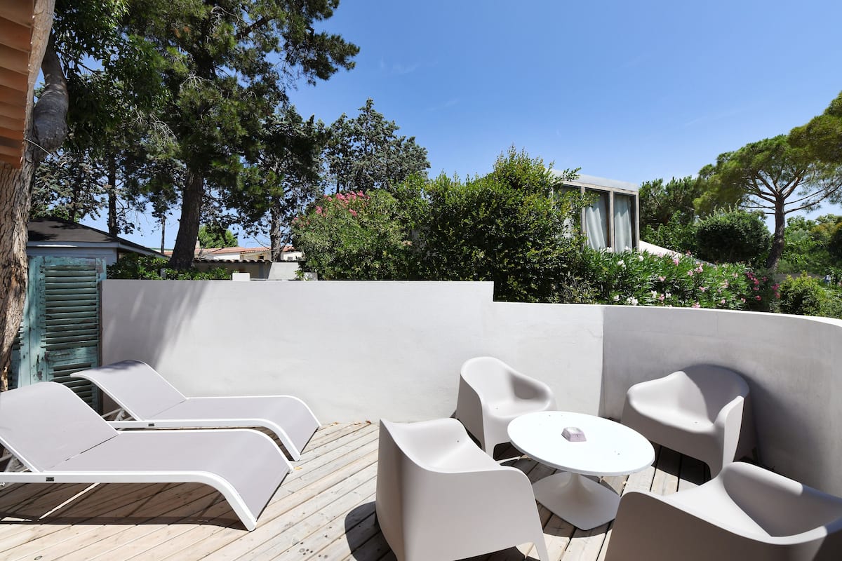 A deck with white lounge chairs and a small round table, surrounded by a white wall and trees.