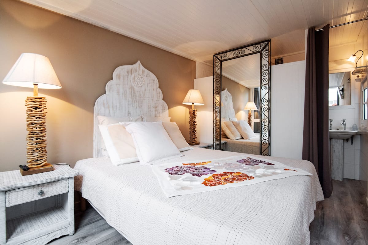 A neatly arranged bedroom with a large bed, two bedside tables, a mirror, and a bathroom visible in the background.